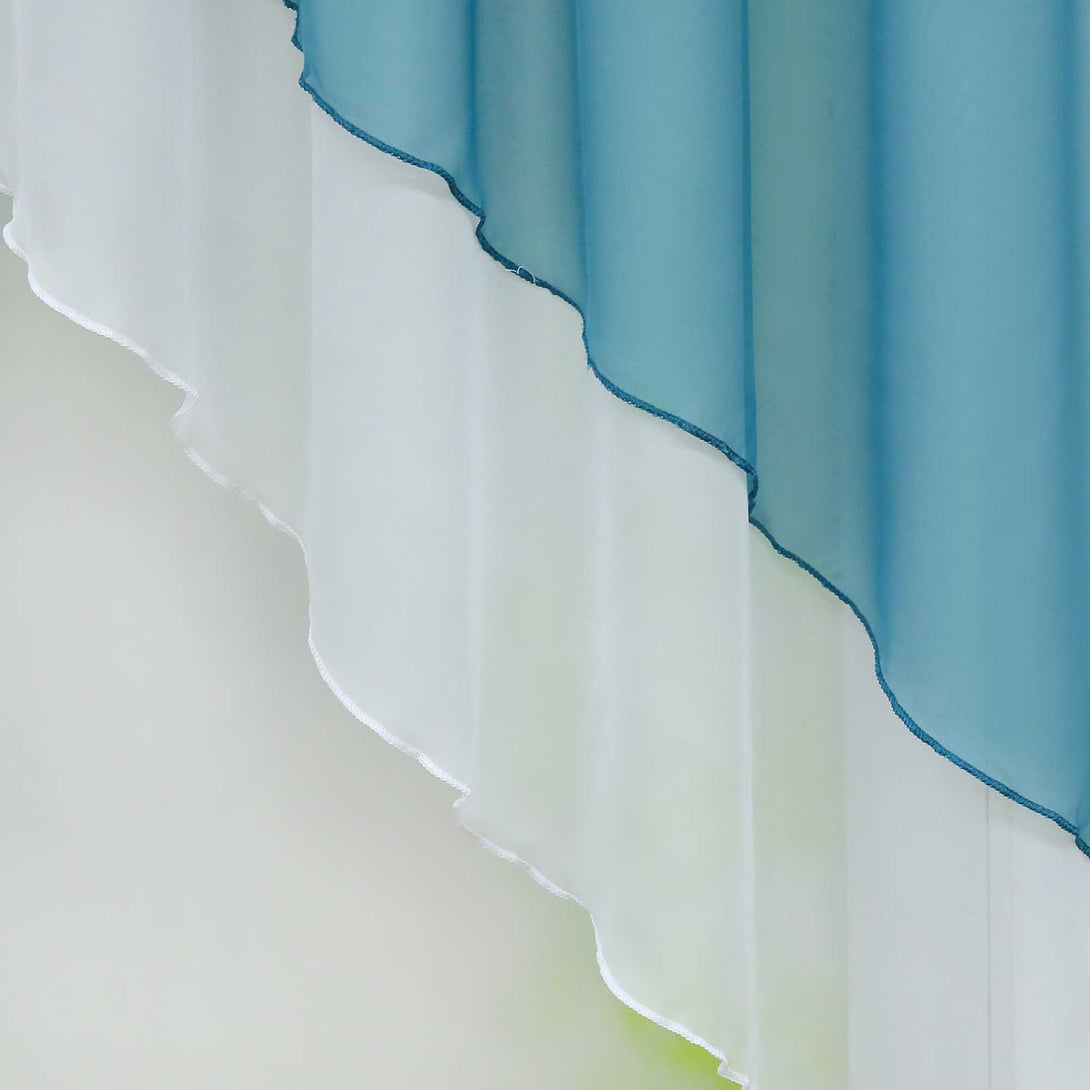Nahaufnahme eines blauen halbtransparenten Gardinenstoffs mit gewellten, sanft fallenden Kanten. Das Licht hebt die feine Struktur des Stoffes hervor, während der Hintergrund in einem sanften Grünton verschwimmt.