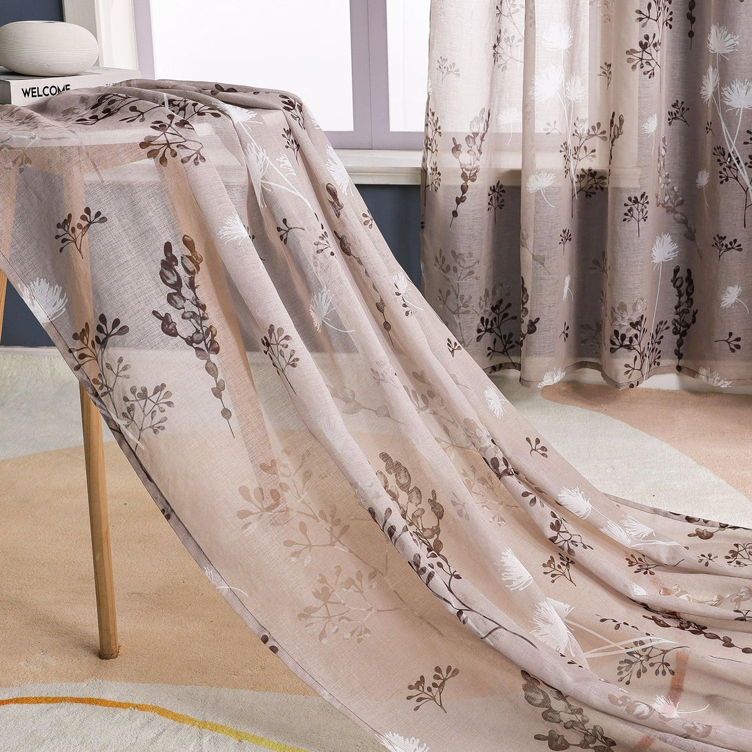 Das Bild zeigt einen transparenten Vorhang mit einem Blumenmuster auf einem hellbeigen Hintergrund, verziert mit weißen Löwenzahnblumen und tiefbraunen Zweigen und Blättern. Ein Teil des Vorhangs hängt über einem Tisch mit Holzbeinen, auf dem weiße Wollbälle und andere Gegenstände liegen. Im Hintergrund sind das Fenster und ein weiterer Teil des Vorhangs zu sehen.