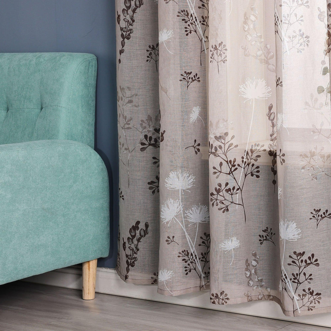 Es zeigt eine Ecke eines Raumes mit einer blauen Wand, vor der ein transparenter Vorhang mit einem Blumenmuster aus weißen Löwenzahnblumen und tiefbraunen Zweigen und Blättern hängt. Daneben steht ein hellgrünes Sofa mit Holzbeinen.