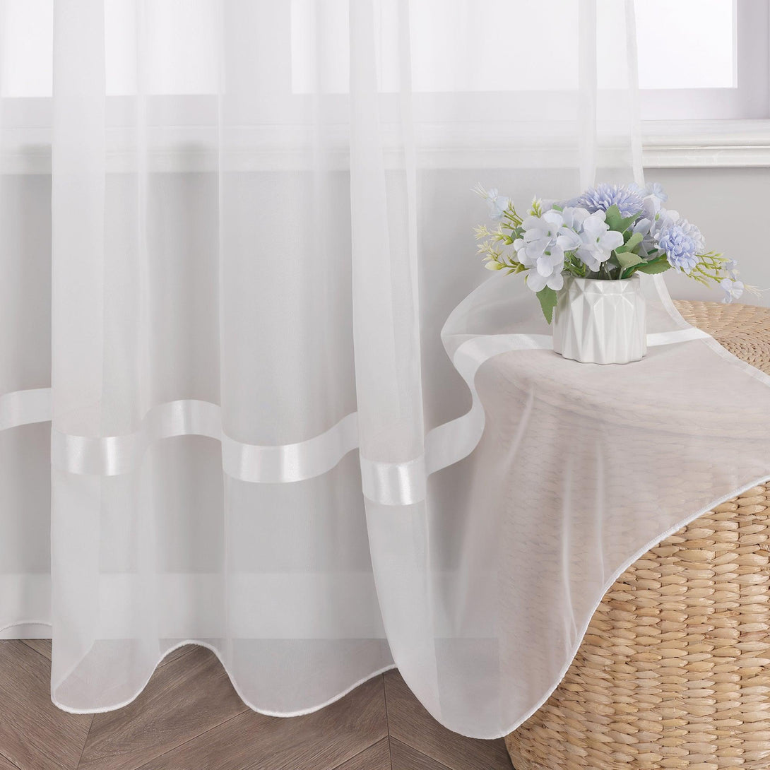 Am Fenster haengt ein weisser Vorhang aus Voile, auf dem sich mehrere weisse Dekobaender befinden. Rechts neben dem Fenster steht ein Objekt aus Flechtmaterial, das mit einem hellbeigen Stoff bedeckt ist. Darauf steht eine weisse Vase mit einem Strauss aus blau-weissen Blumen.