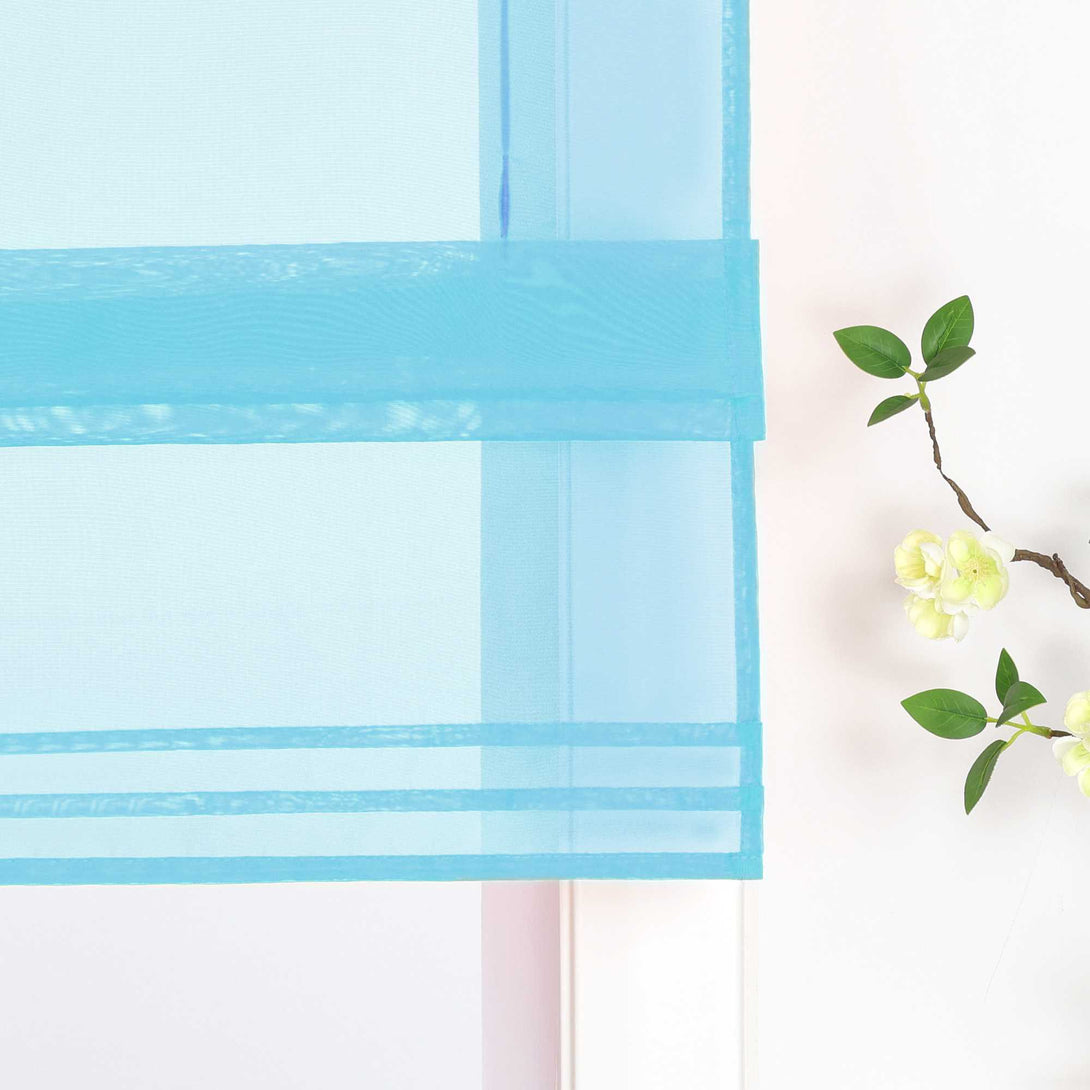 Detailansicht des unteren Bereichs eines halbtransparenten blauen Raffrollos. Der Stoff ist sanft hochgezogen und bildet dekorative Falten. Eine stilvolle Blumen-Deko neben dem Fenster rundet das Gesamtbild harmonisch ab.