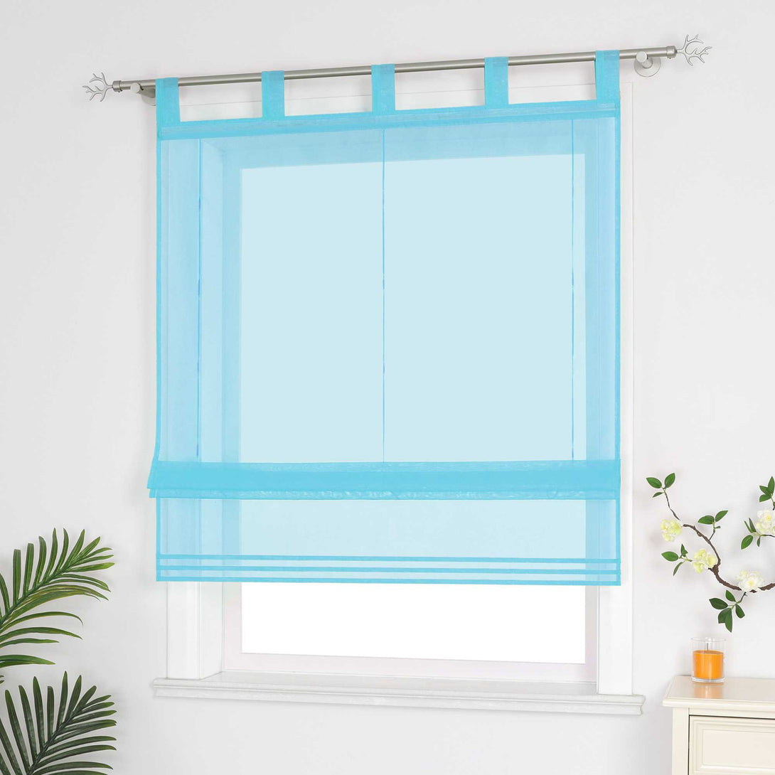 Halbtransparenter blauer Raffrollo hängt vor dem Fenster und sorgt für eine helle, luftige Atmosphäre. Daneben setzen grüne Pflanzen dekorative Akzente. Seitliche Ansicht.