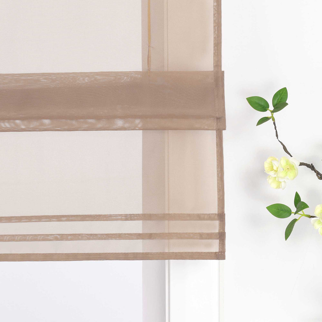 Detailansicht des unteren Bereichs eines halbtransparenten braunen Raffrollos. Der Stoff ist sanft hochgezogen und bildet dekorative Falten. Eine stilvolle Blumen-Deko neben dem Fenster rundet das Gesamtbild harmonisch ab.