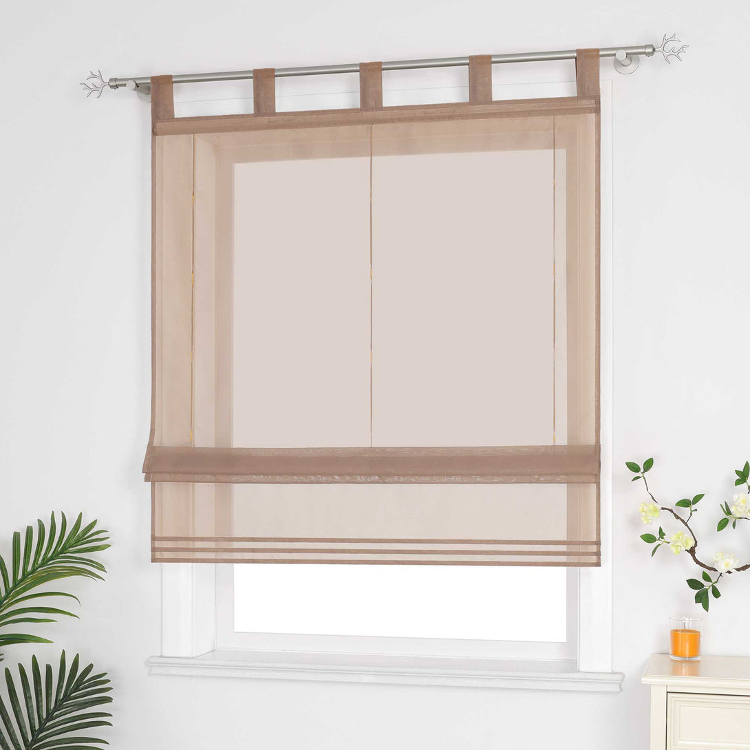 Halbtransparenter brauner Raffrollo hängt vor dem Fenster und sorgt für eine helle, luftige Atmosphäre. Daneben setzen grüne Pflanzen dekorative Akzente. Seitliche Ansicht.