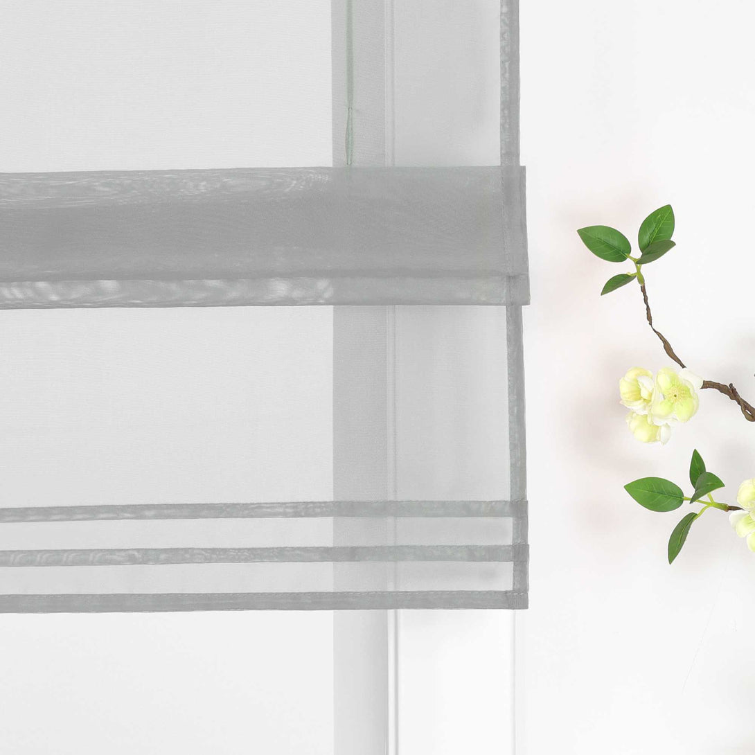 Detailansicht des unteren Bereichs eines halbtransparenten grauen Raffrollos. Der Stoff ist sanft hochgezogen und bildet dekorative Falten. Eine stilvolle Blumen-Deko neben dem Fenster rundet das Gesamtbild harmonisch ab.