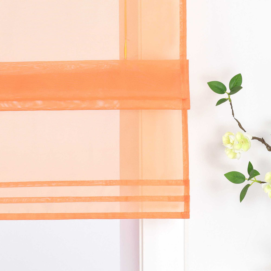 Detailansicht des unteren Bereichs eines halbtransparenten orangen Raffrollos. Der Stoff ist sanft hochgezogen und bildet dekorative Falten. Eine stilvolle Blumen-Deko neben dem Fenster rundet das Gesamtbild harmonisch ab.