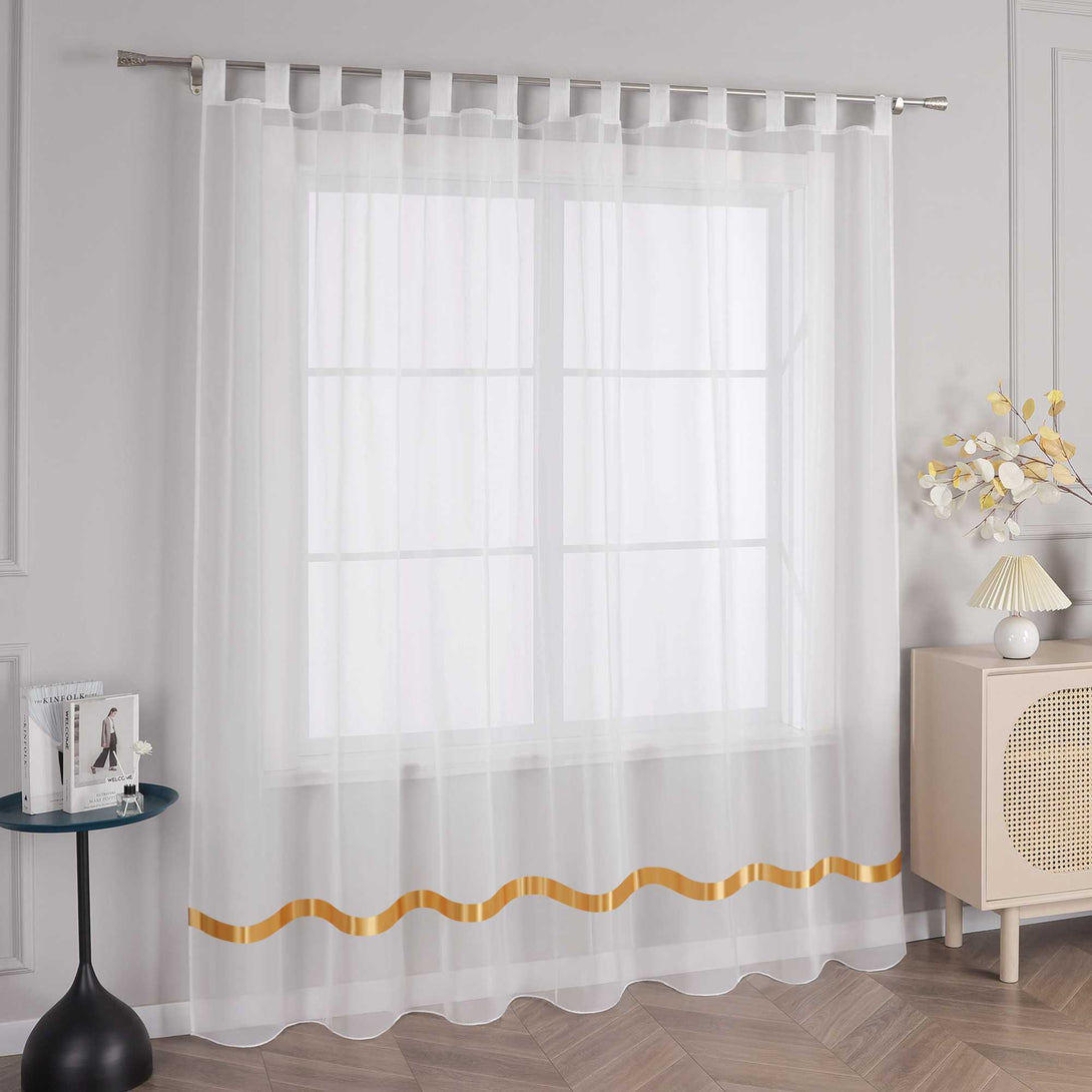 An einem Fenster haengt ein weisser Vorhang mit orangefarbenen Satinbaendern und Schlaufen. Am oberen Ende fuehren die Schlaufen durch eine Metallstange, um den Vorhang an seinem Platz zu halten. Links vom Fenster steht ein kleiner schwarzer runder Tisch mit einigen Gegenstaenden. Rechts daneben befindet sich ein heller Schrank mit einer Lampe mit hellem Schirm und einem Strauss dekorativer Blumen.