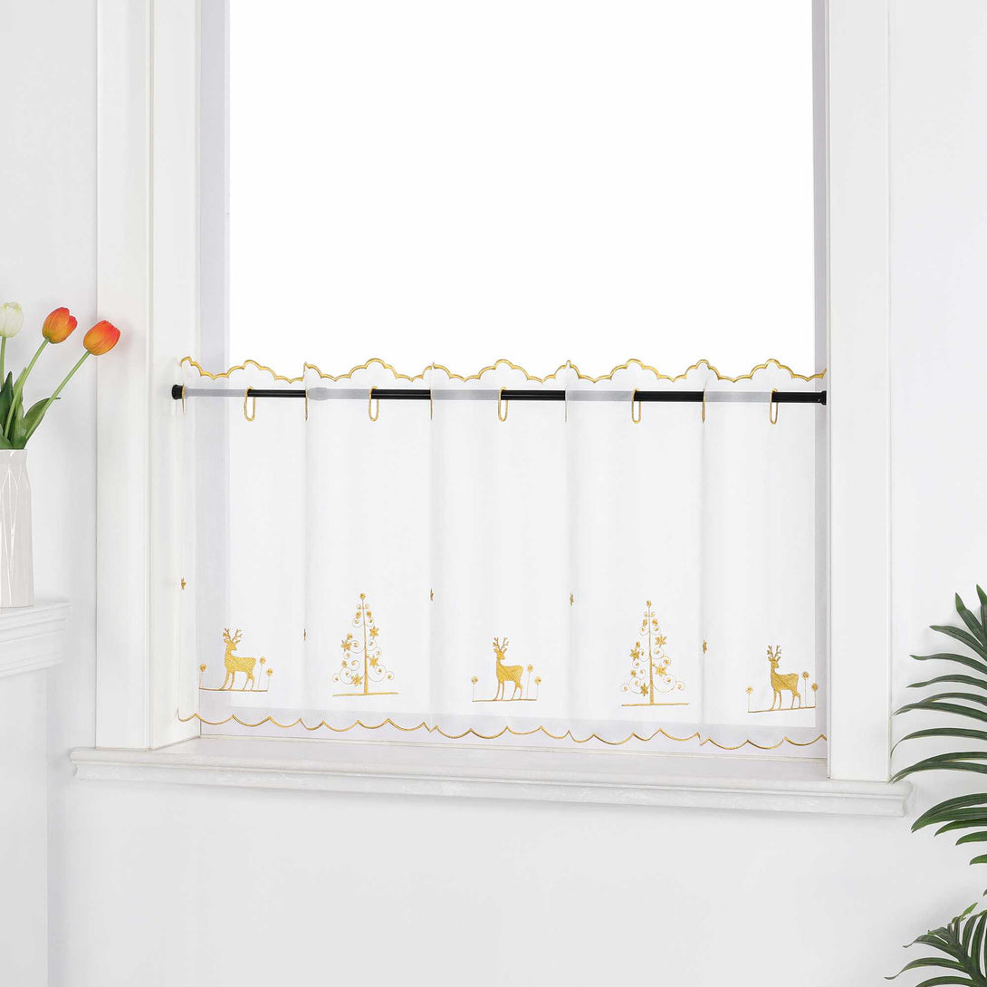 Die transparente Kurzgardine hängt festlich vor einem weißen Fenster. Die Gardine ist mit goldenen Nähten verziert und zeigt handgestickte Motive von Schneeflocken, Rentieren und einem Weihnachtsbaum, die eine weihnachtliche Atmosphäre schaffen. Die weiße Wand im Hintergrund sorgt für eine helle und neutrale Kulisse, während die pflanzlichen Dekorationen rund um das Fenster eine natürliche und gemütliche Stimmung verleihen. Der transparente Stoff lässt viel Tageslicht herein und sorgt für eine subtile, aber