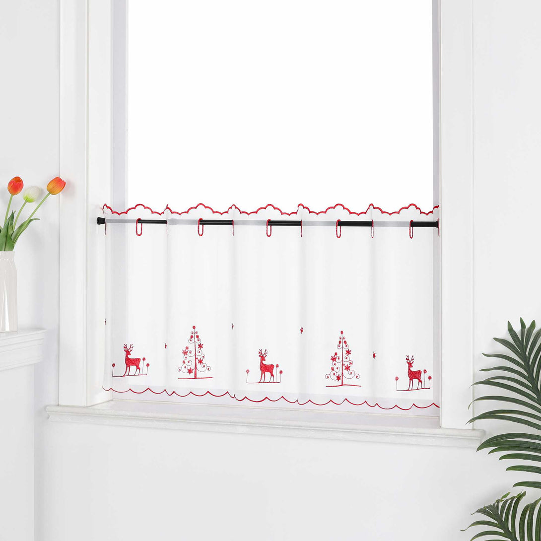 Die transparente Kurzgardine hängt festlich vor einem weißen Fenster. Die Gardine ist mit roten Nähten verziert und zeigt handgestickte Motive von Schneeflocken, Rentieren und einem Weihnachtsbaum, die eine weihnachtliche Atmosphäre schaffen. Die weiße Wand im Hintergrund sorgt für eine helle und neutrale Kulisse, während die pflanzlichen Dekorationen rund um das Fenster eine natürliche und gemütliche Stimmung verleihen. Der transparente Stoff lässt viel Tageslicht herein und sorgt für eine subtile, aber fe