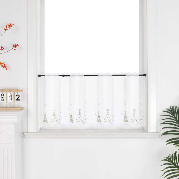 Weiße, transparente Scheibengardine mit goldenen Weihnachtsbaum-Motiven hängt elegant vor einem weißen Fensterrahmen. Die Gardine lässt sanftes Licht herein und schafft eine festliche Atmosphäre. Weiße Wände und dekorative Blumenarrangements rund um das Fenster unterstreichen den hellen, freundlichen Stil.