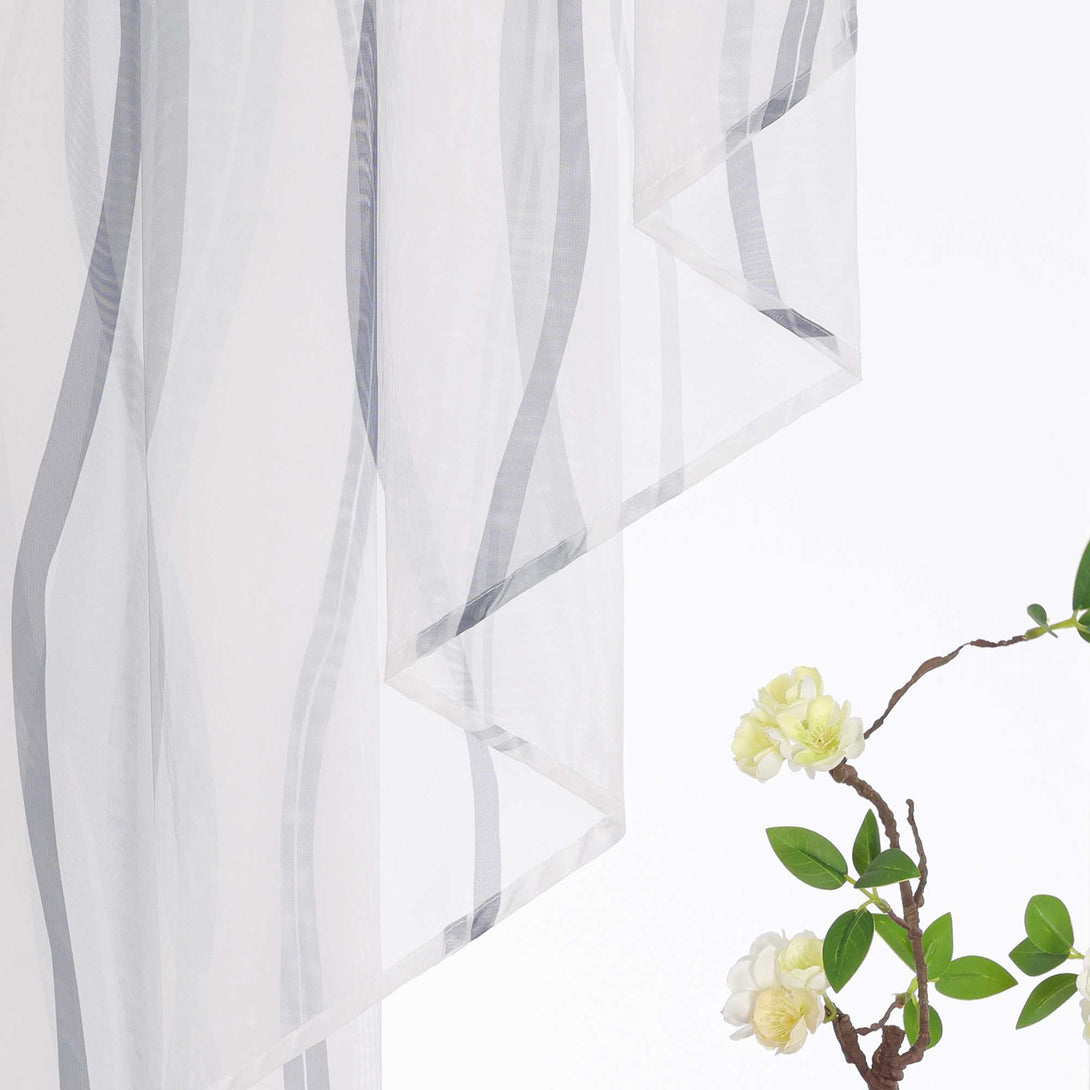 Das Bild zeigt einen weißen Voile-Vorhang mit einem grauen Linienmuster, daneben befindet sich ein Zweig mit grünen Blättern und gelb-weißen Blüten. Der Voile-Vorhang ist zart und lichtdurchlässig, während die grauen Linien dem Design eine künstlerische Note verleihen und eine stilvolle Atmosphäre schaffen.