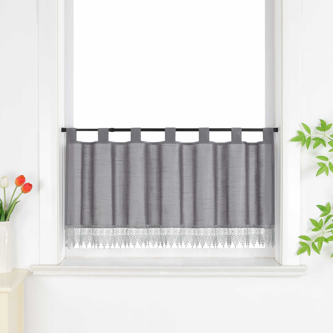 Graue Scheibengardine hängt vor einem Fenster mit weißer Wand. Daneben sorgen rote Blumen, grüne Blätter und eine Vase für eine frische und dekorative Note.