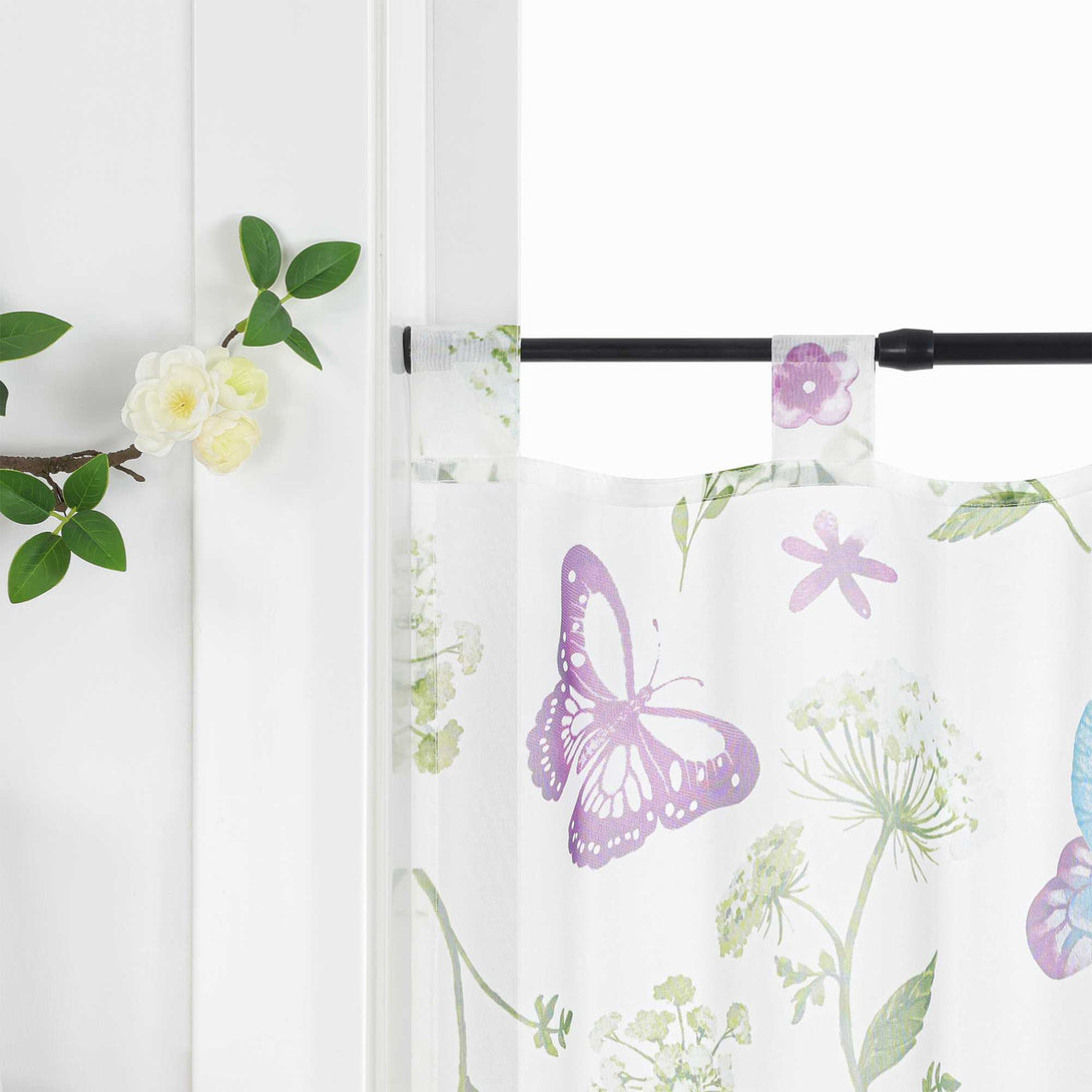 Eine weiße Scheibengardine mit lila Schmetterlingen, grünen Zweigen und weißen Blumen, aufgehängt an einem schwarzen Stab. Auf der linken Seite der Gardine an der weißen Wand befindet sich ein Ast mit grünen Blättern und weißen Blüten.