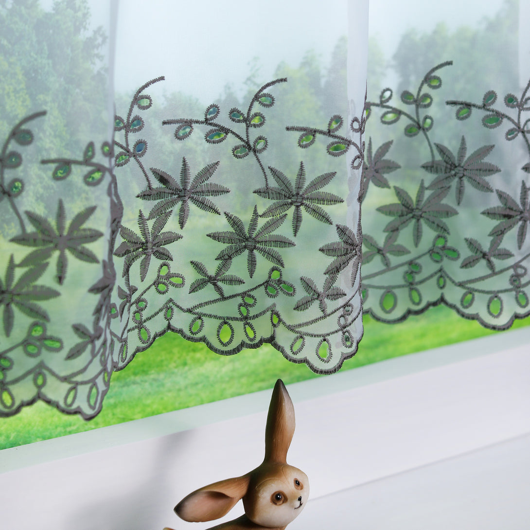 Ein bestickter, halbtransparenter Vorhang mit schwarzen Ahornblatt-Motiven. Der untere Rand hat ein elegantes, wellenförmiges Finish. Eine Hasenfigur ziert die Fensterbank darunter.