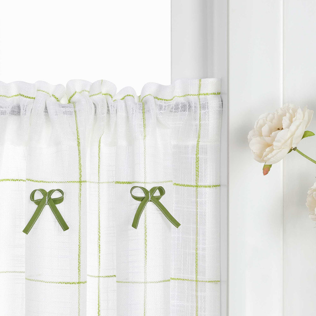 Dies ist ein Ausschnitt von Gardinen im Landhausstil. Auf dem weißen Stoff befindet sich ein Gitternmuster aus hellgrünen Linien, in dem grüne Schleifen als Dekoration verteilt sind.
