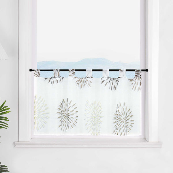 Die transparente Scheibengardine mit braunen Feuerwerksmotiven hängt elegant vor einem Fenster. Die weiße Wand im Hintergrund lässt die braunen Muster des Feuerwerks besonders hervorstechen und sorgt für eine ruhige, festliche Atmosphäre im Raum.