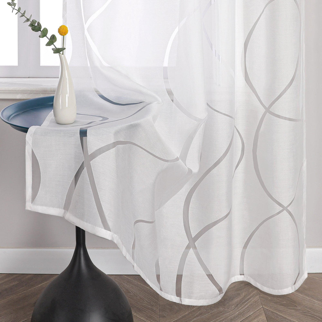Das Bild zeigt einen weißen, transparenten Voile-Vorhang mit einem kurvigen Muster. Der Stoff ist leicht und lichtdurchlässig. Eine Ecke des Vorhangs liegt auf einem schwarzen runden Tisch, auf dem eine Vase mit gelben Blumen steht.
