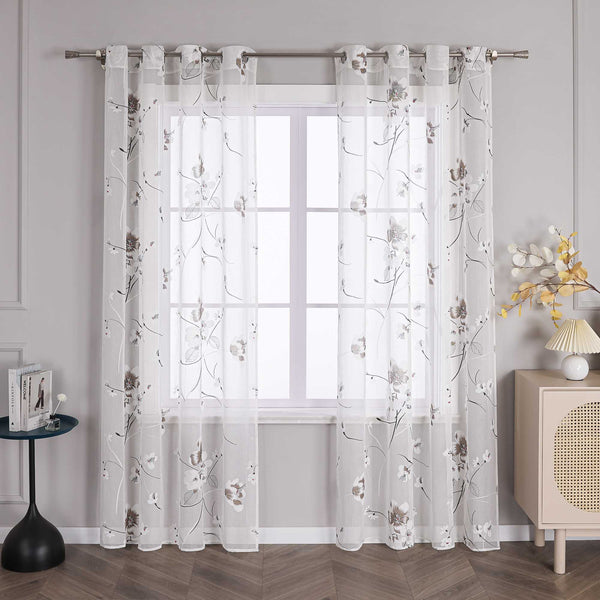 Das Bild zeigt ein Fenster, an dem ein Paar weiße Gardinen hängt. Die Gardinen sind mit einem filigranen Blumenmuster verziert, wobei die Blüten hauptsächlich in Weiß und Braun gehalten sind. Die Blütenmitte ist mit roten Akzenten verziert, und die Zweige sind in schwarzen Linien dargestellt. Die Gardinen sind an einer Metallstange befestigt, die an beiden Enden an einer hellgrauen Wand montiert ist.