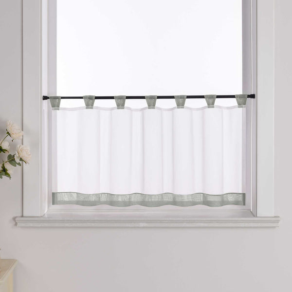 Die graue Scheibengardine hängt vor einem Fenster, wobei die Hintergrundwand weiß ist. Der Vorhang ist transparent und wird von blumigen Dekorationen an der Seite begleitet.