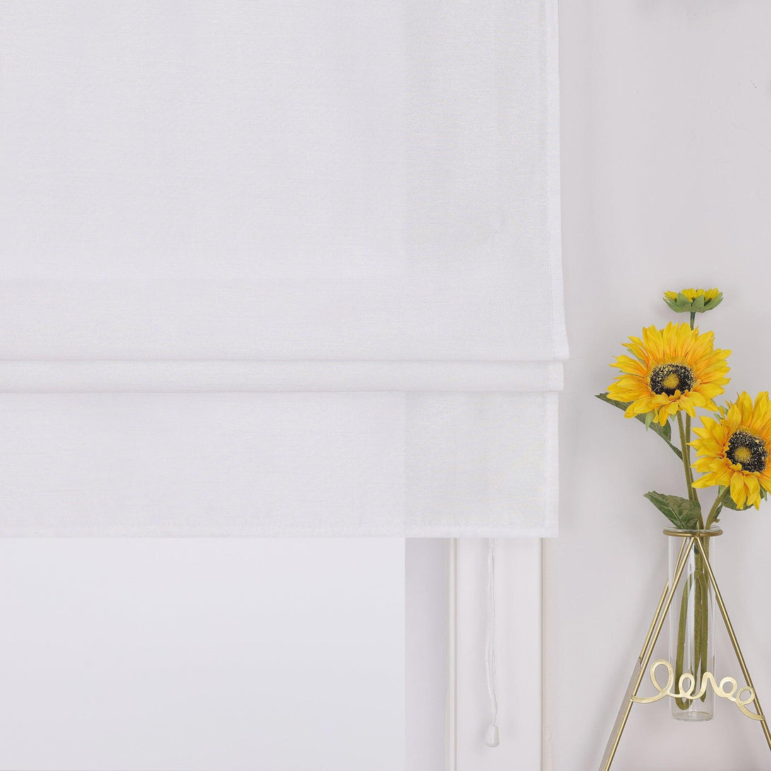 Das Bild zeigt ein Detail einer weißen Raffrollo, der sanft am Fenster hängt. Neben dem Vorhang steht eine transparente Glasvase mit gelben Sonnenblumen, die in einem goldenen Halter arrangiert sind, was dem Raum eine warme, einladende Atmosphäre verleiht.